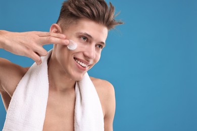Handsome man applying moisturizing cream onto his face on blue background. Space for text