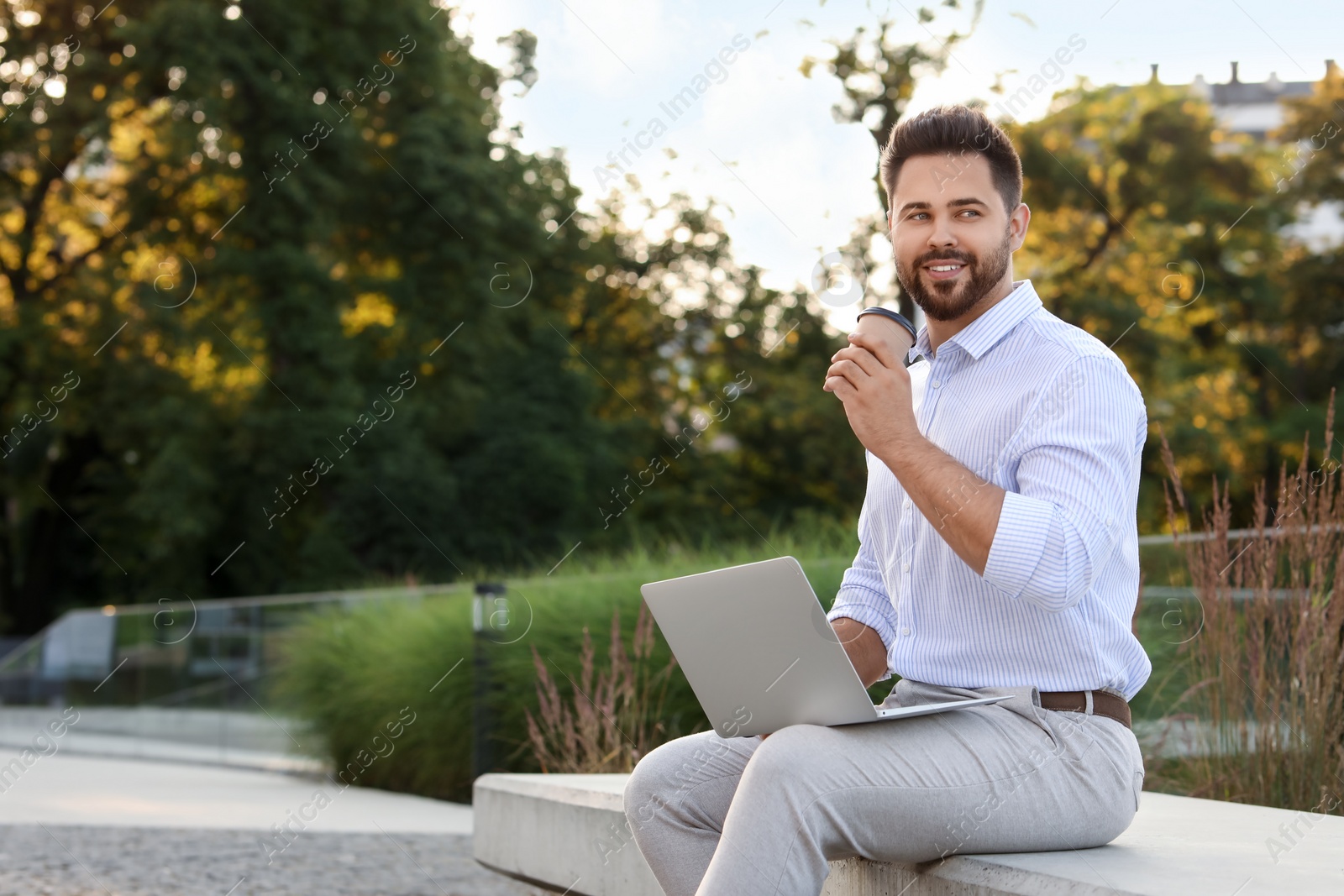 Photo of Handsome young man with cup of coffee using laptop on stone bench outdoors. Space for text