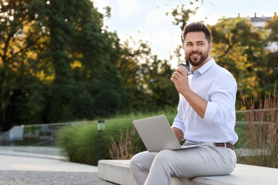 Photo of Handsome young man with cup of coffee using laptop on stone bench outdoors. Space for text