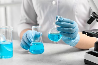 Photo of Scientist with flasks of light blue liquid at white table in laboratory, closeup