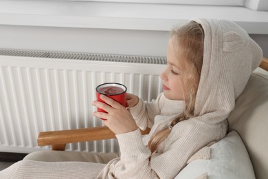Photo of Little girl with cup of hot drink near heating radiator indoors
