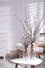 Glass vases with pussy willow tree branches on table near armchair in room