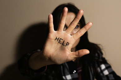 Abused young woman showing palm with word HELP near beige wall, focus on hand. Domestic violence concept