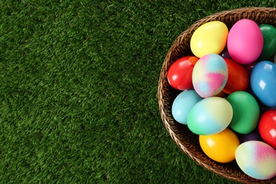 Photo of Wicker basket with bright painted Easter eggs on green grass, top view. Space for text