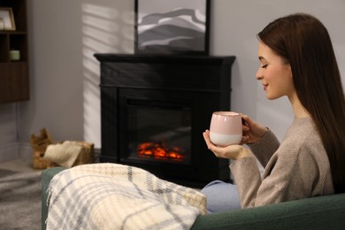 Beautiful young woman with cup of hot drink sitting on sofa near fireplace. Space for text