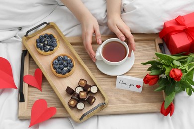 Photo of Tasty breakfast served in bed. Woman with tea, desserts, gift box, flowers and I Love You card at home, above view