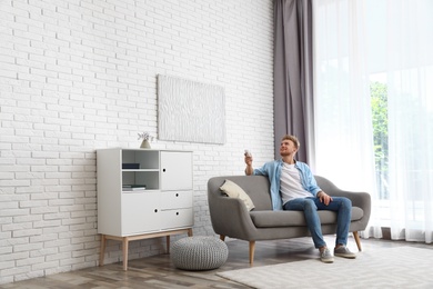 Photo of Happy young man switching on air conditioner on sofa at home