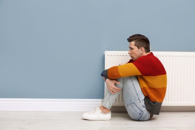 Photo of Sad young man suffering from cold on floor near radiator