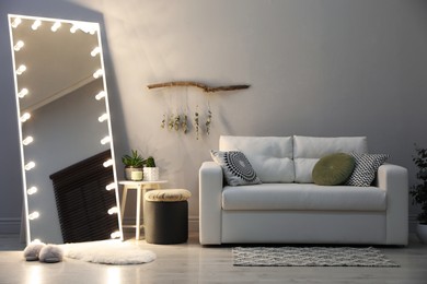 Stylish mirror with light bulbs and comfortable sofa in living room. Interior design