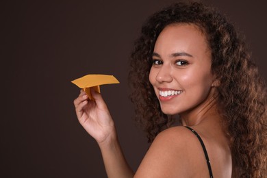 Beautiful African-American woman playing with paper plane on brown background