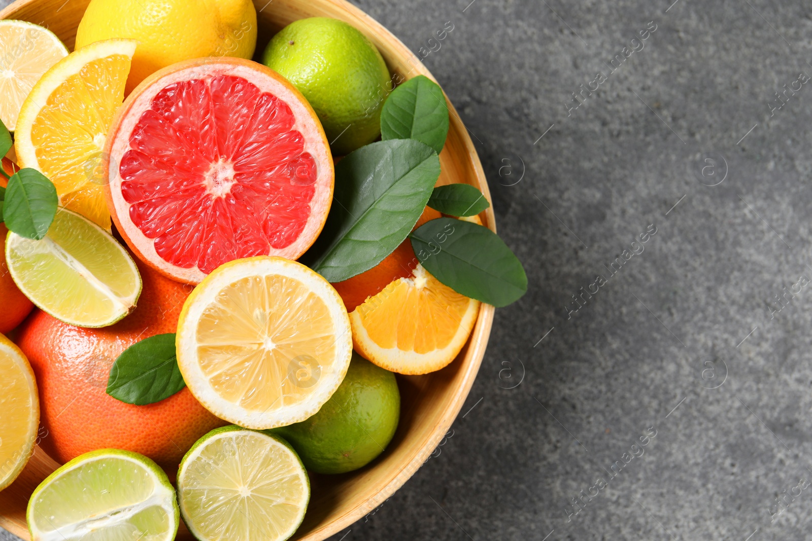 Photo of Different fresh citrus fruits and leaves in bowl on grey textured table, top view. Space for text