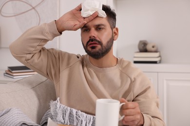 Photo of Sick man with tissue and cup of drink on sofa at home. Cold symptoms