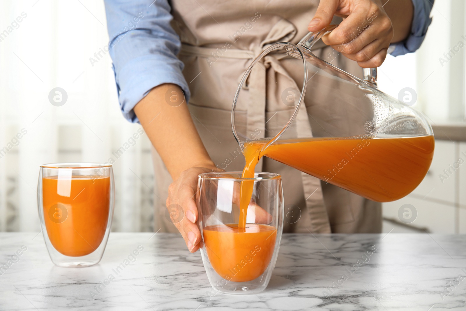 Photo of Woman pouring freshly made carrot juice into glass on white marble table in kitchen, closeup
