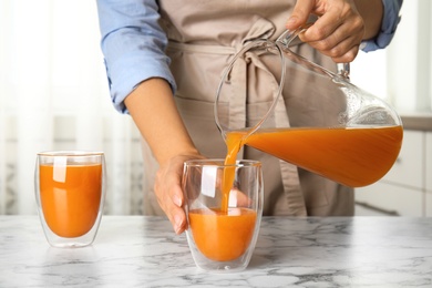 Woman pouring freshly made carrot juice into glass on white marble table in kitchen, closeup