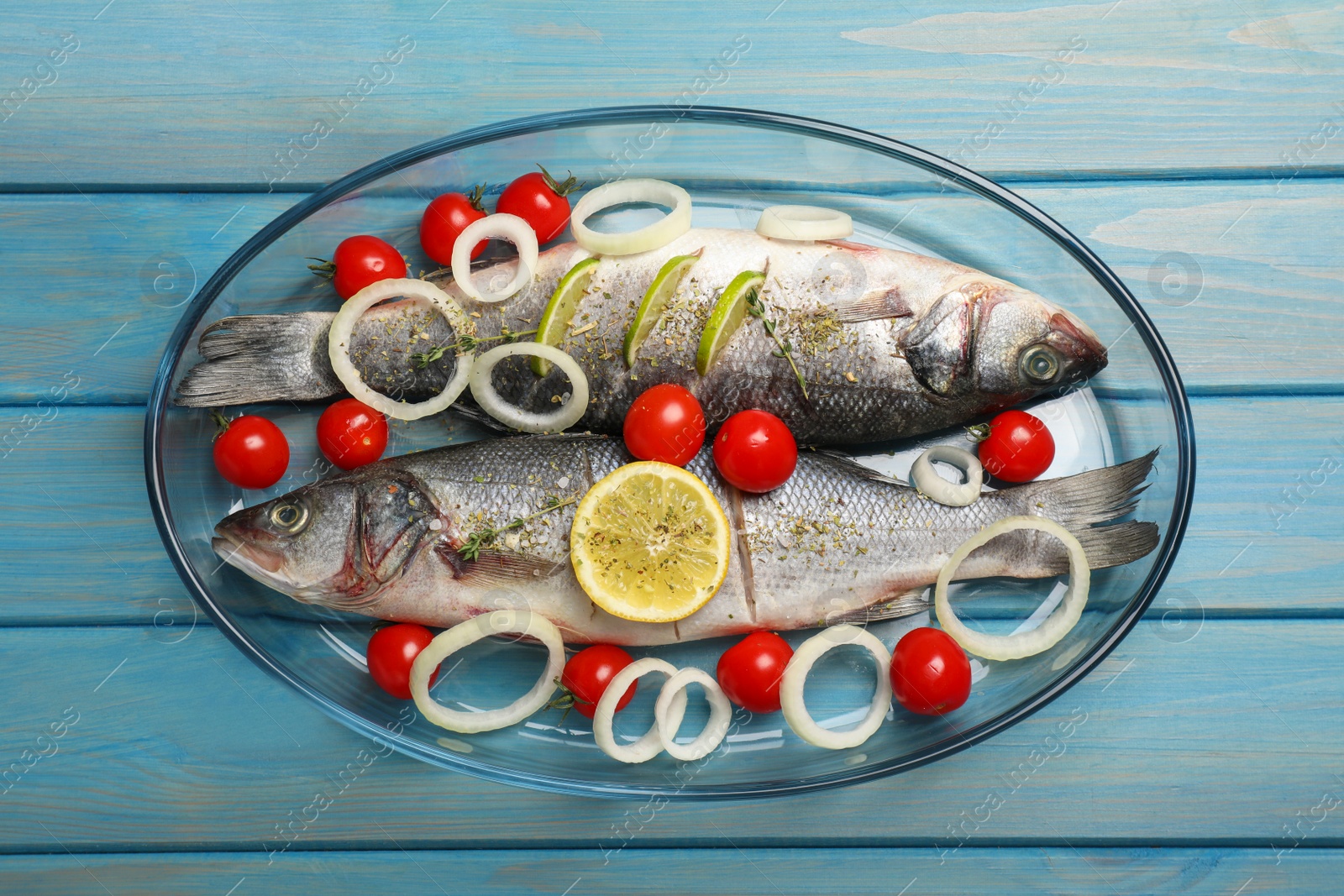 Photo of Glass baking tray with sea bass fish and ingredients on light blue wooden table, top view