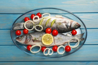 Photo of Glass baking tray with sea bass fish and ingredients on light blue wooden table, top view