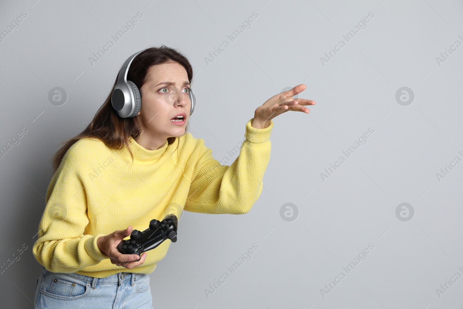 Photo of Confused woman in headphones with game controller on light grey background. Space for text