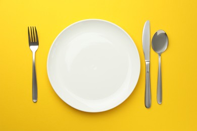 Photo of Clean plate, fork, knife and spoon on yellow background, flat lay