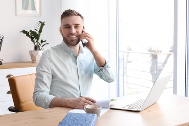 Young man talking on mobile phone while working with laptop at desk. Home office