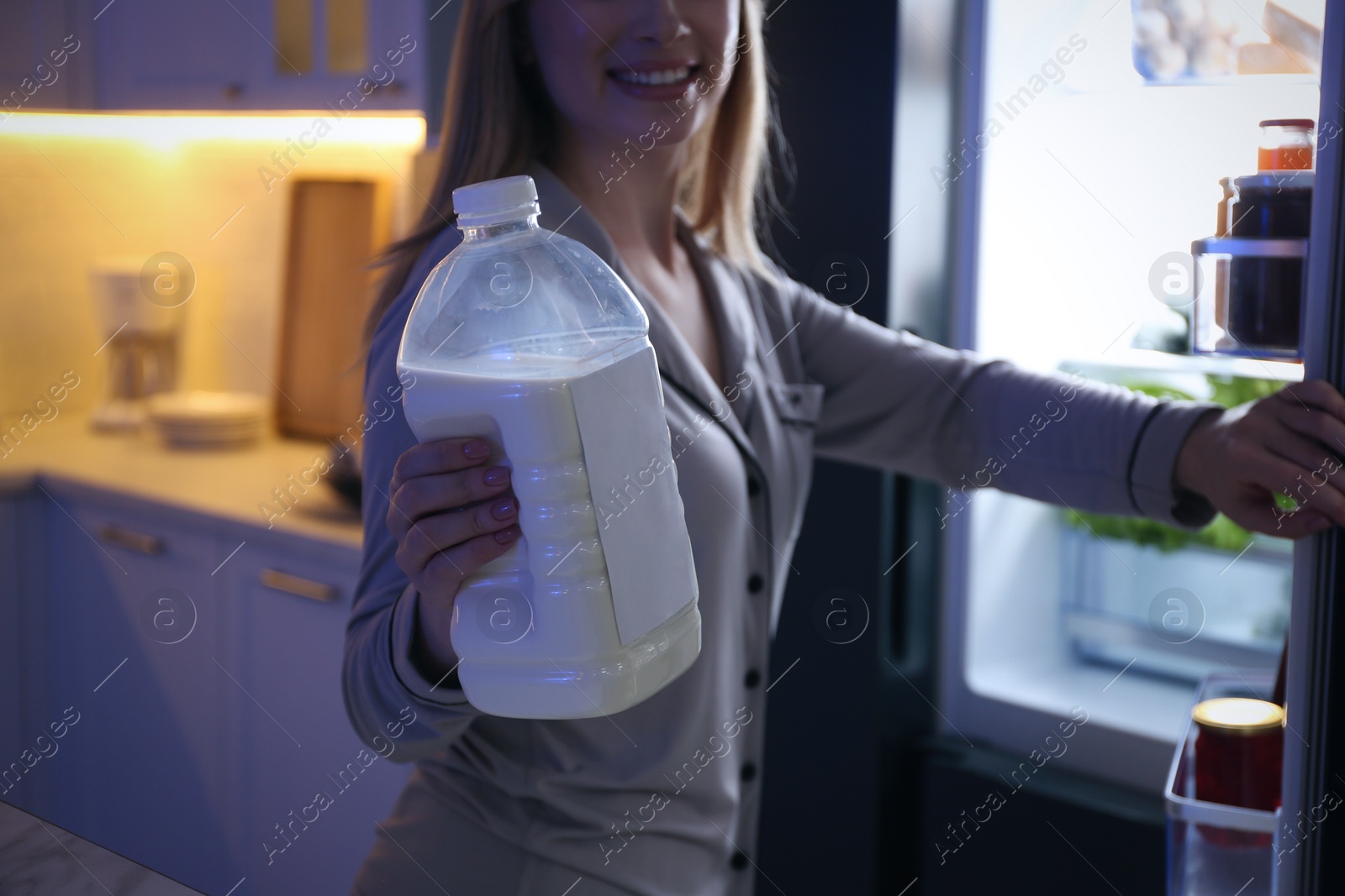 Photo of Young woman holding gallon bottle of milk near refrigerator in kitchen at night, closeup