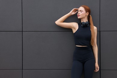 Photo of Beautiful woman in stylish gym clothes with headphones near dark grey wall on street, space for text