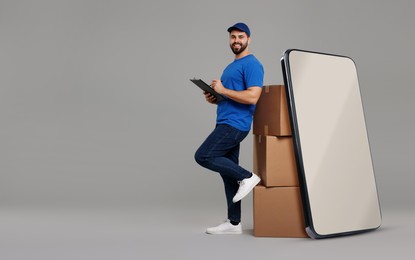 Courier with stack of parcels and clipboard near huge smartphone on grey background. Delivery service. Space for text