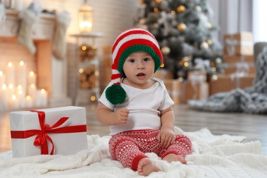 Cute little baby with elf hat near Christmas gift on floor at home