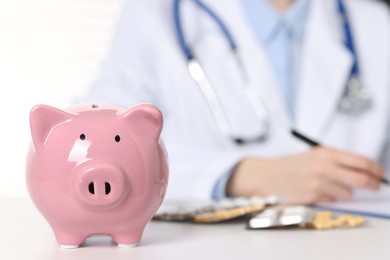 Photo of Doctor making notes at white table, focus on piggy bank