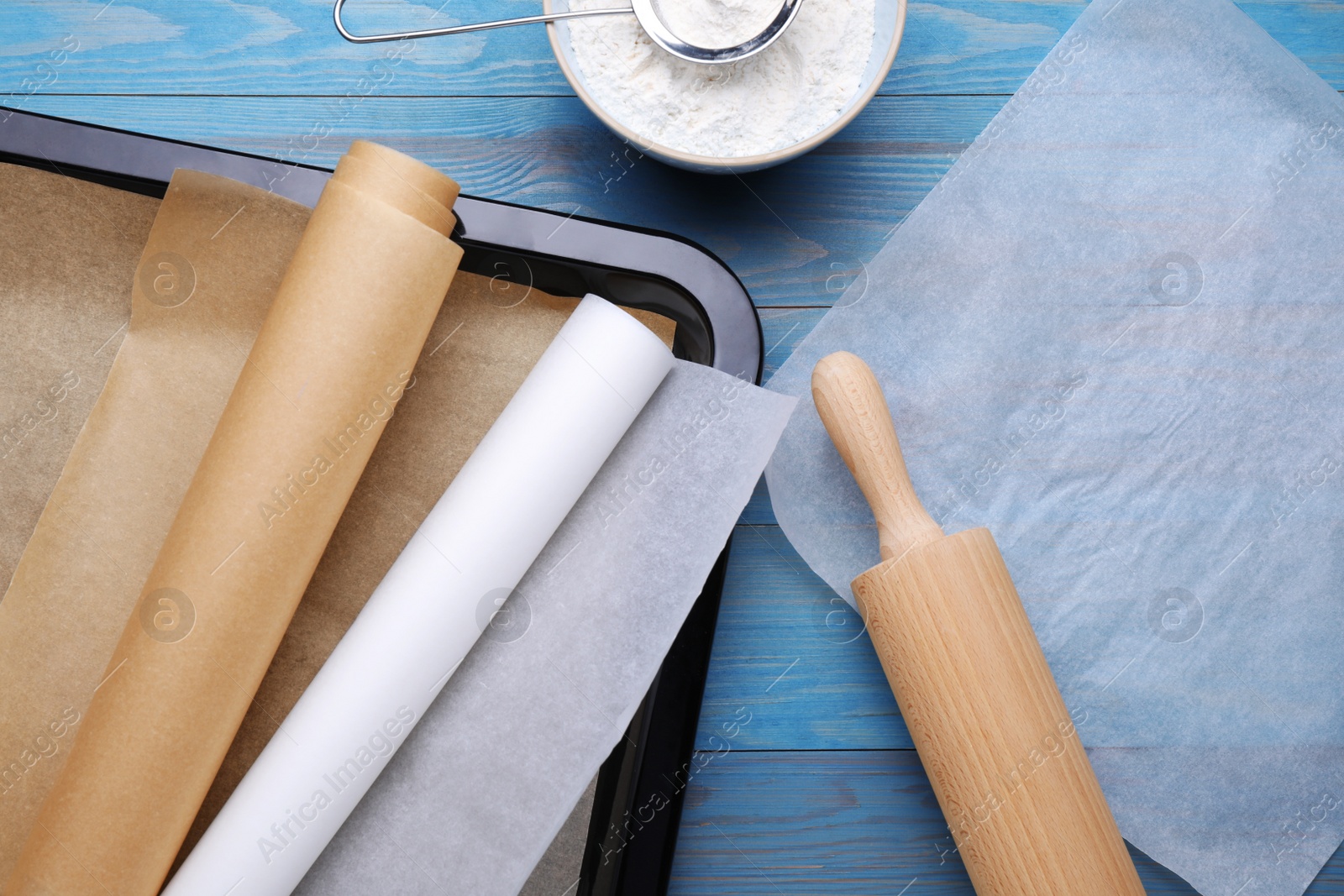 Photo of Parchment paper, baking pan, rolling pin and flour on light blue wooden table, flat lay