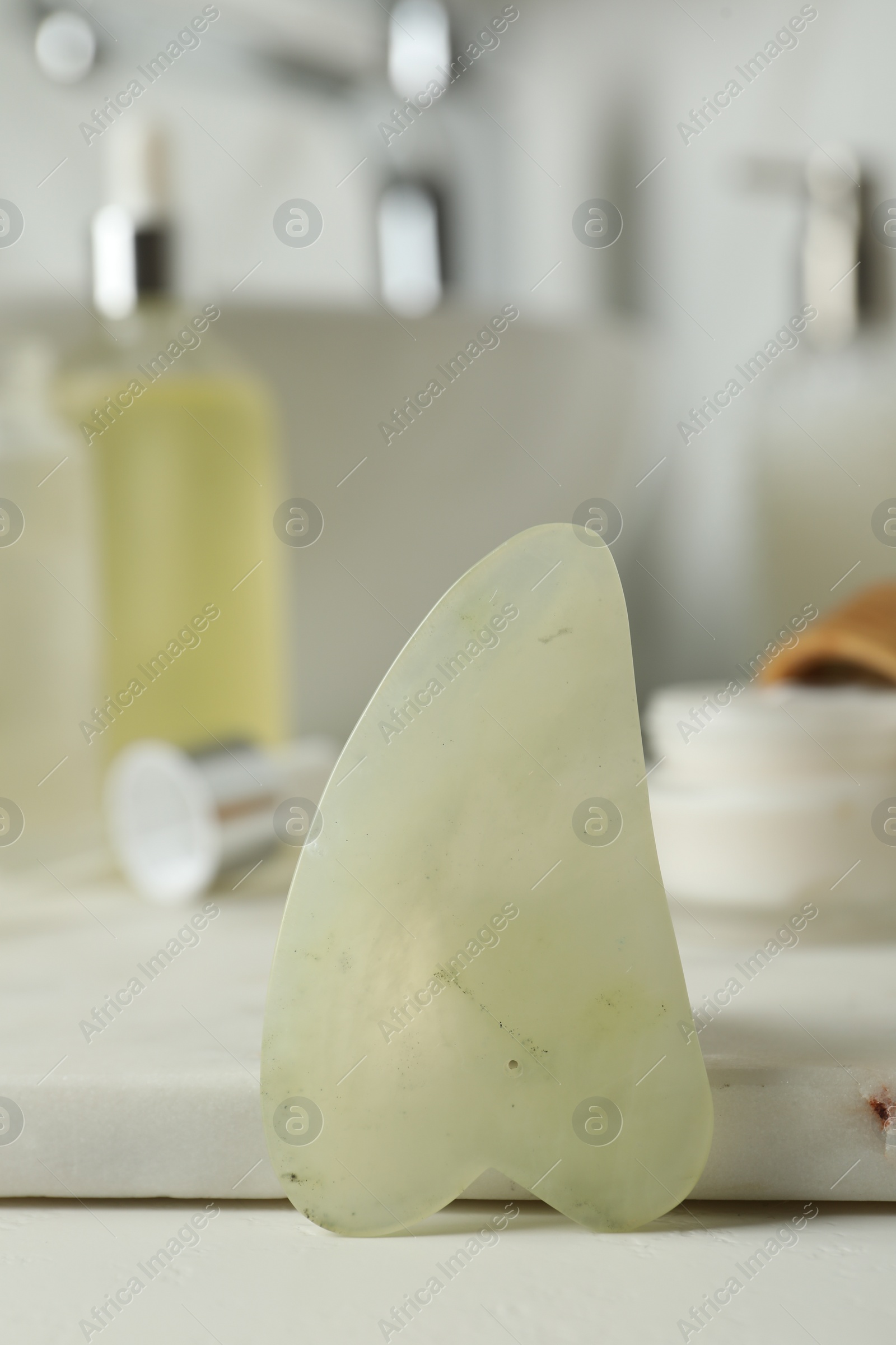 Photo of Jade gua sha tool and toiletries on white countertop in bathroom, closeup