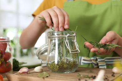 Photo of Woman putting herbs into pickling jar at table in kitchen, closeup