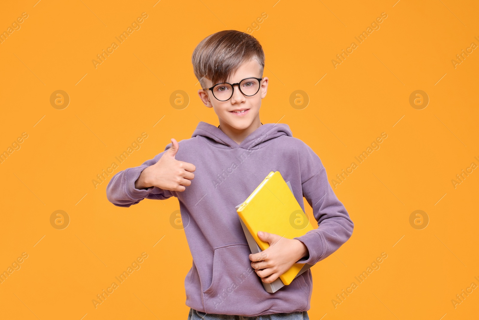 Photo of Cute schoolboy in glasses holding books and showing thumbs up on orange background