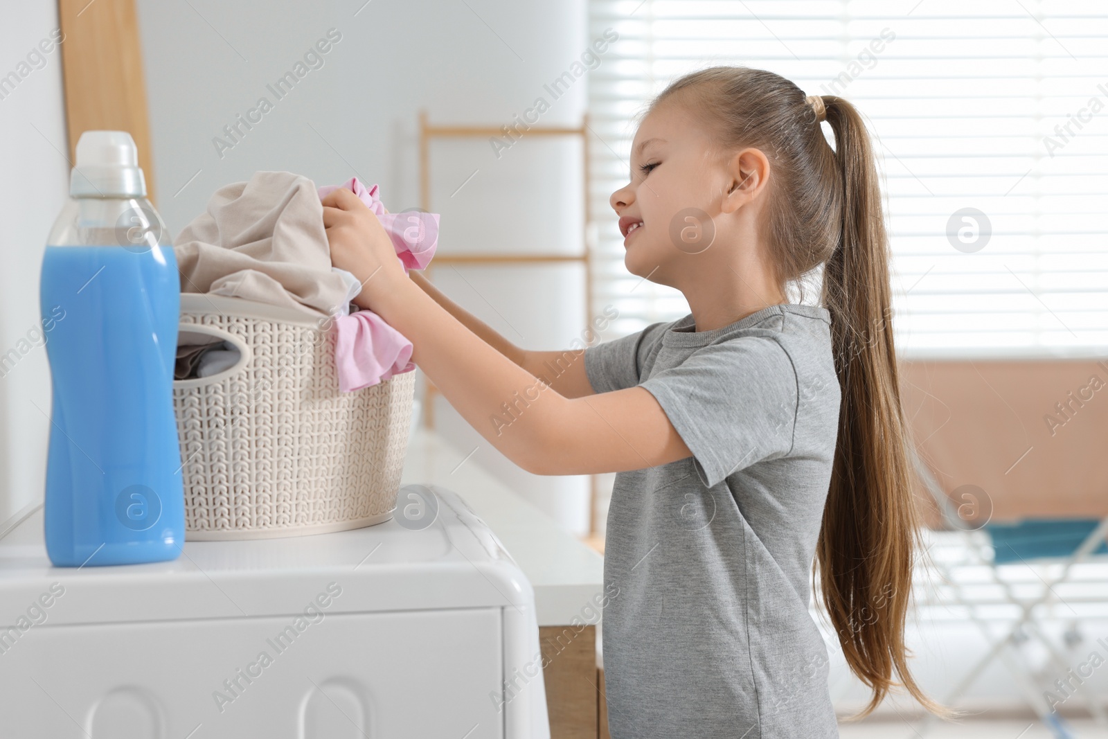 Photo of Little girl taking out dirty clothes from basket in bathroom