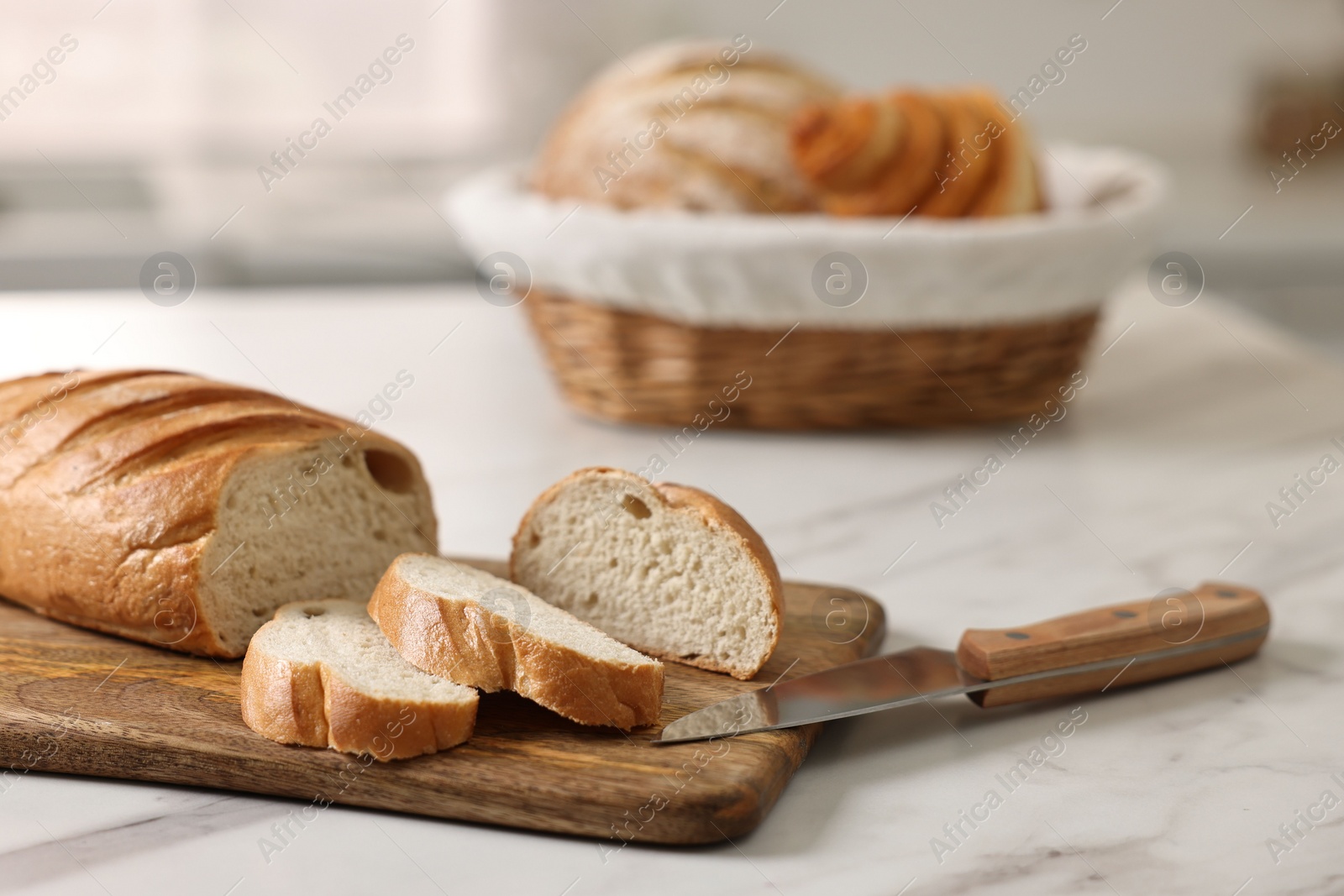 Photo of Wicker bread basket with freshly baked loaves and knife on white marble table in kitchen