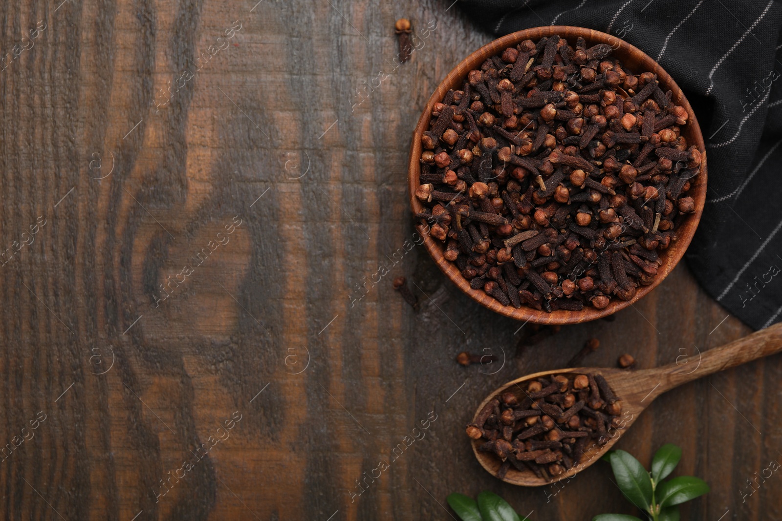 Photo of Aromatic cloves in bowl, spoon and green leaves on wooden table, flat lay. Space for text