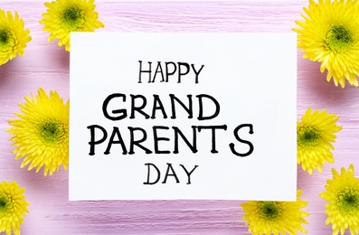 Photo of Beautiful yellow flowers and card with phrase Happy Grandparents Day on pink wooden background, flat lay