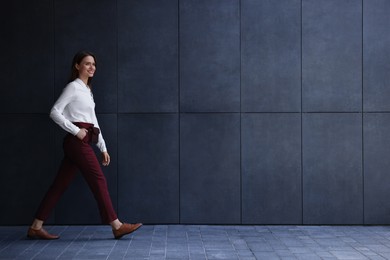 Young woman in formal clothes walking near grey wall outdoors, space for text