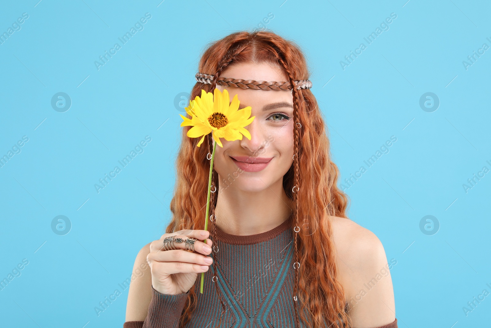 Photo of Beautiful young hippie woman covering eye with sunflower on light blue background