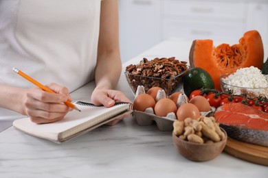 Woman with notebook and healthy food at white table, closeup. Keto diet