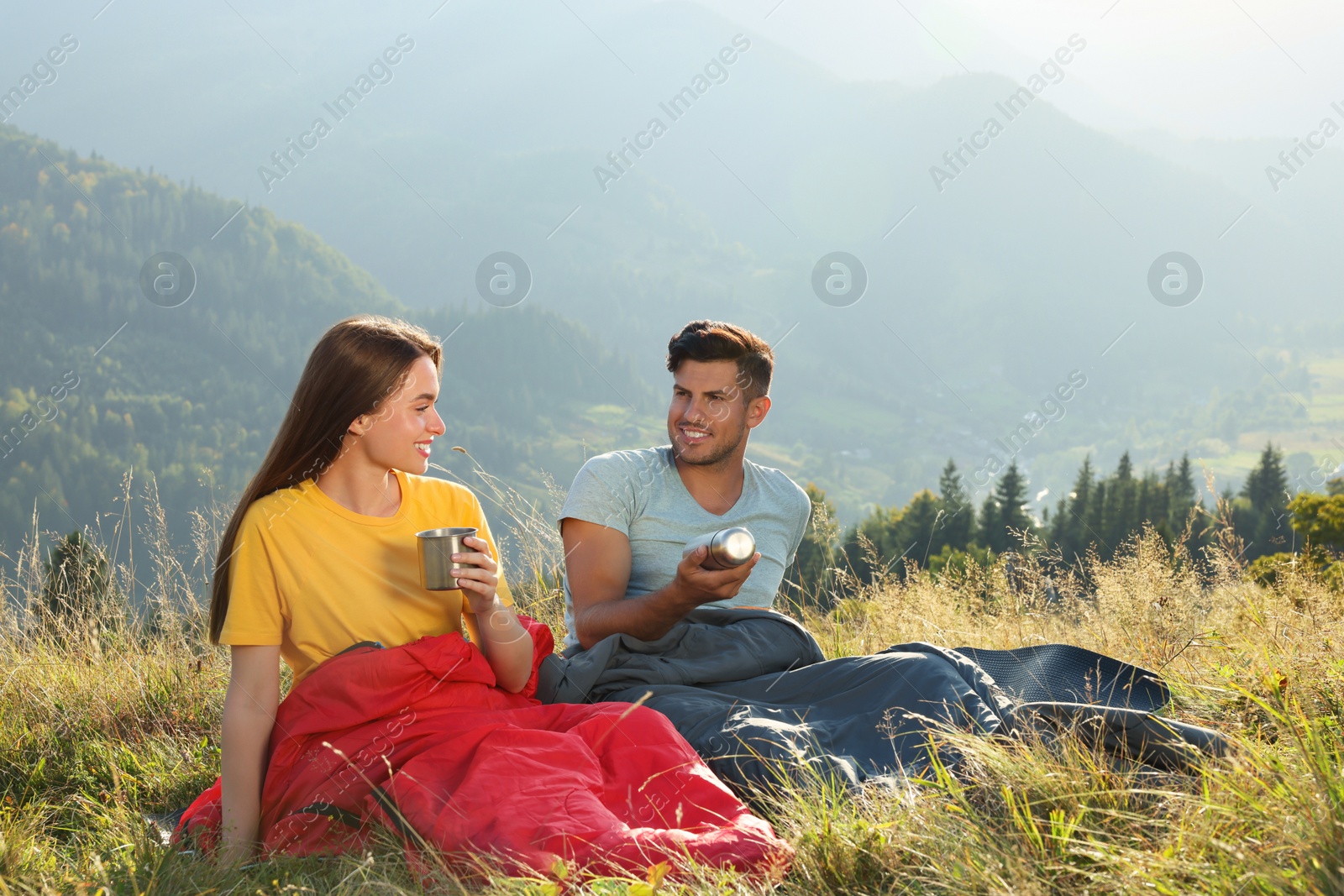 Photo of Happy couple with drinks in sleeping bags surrounded by beautiful nature