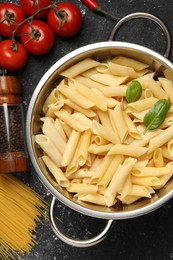 Photo of Cooked pasta in metal colander, products and peppercorns on dark textured table, flat lay