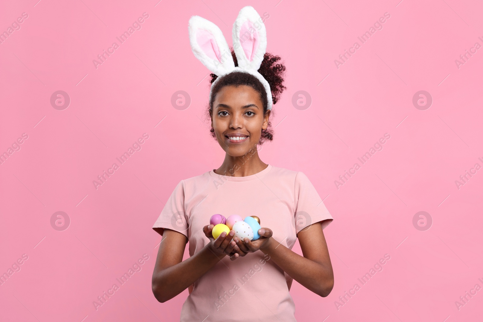 Photo of Happy African American woman in bunny ears headband holding Easter eggs on pink background