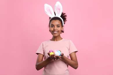 Happy African American woman in bunny ears headband holding Easter eggs on pink background