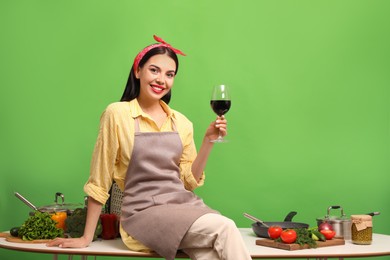 Young housewife with glass of wine, vegetables and different utensils on green background. Space for text
