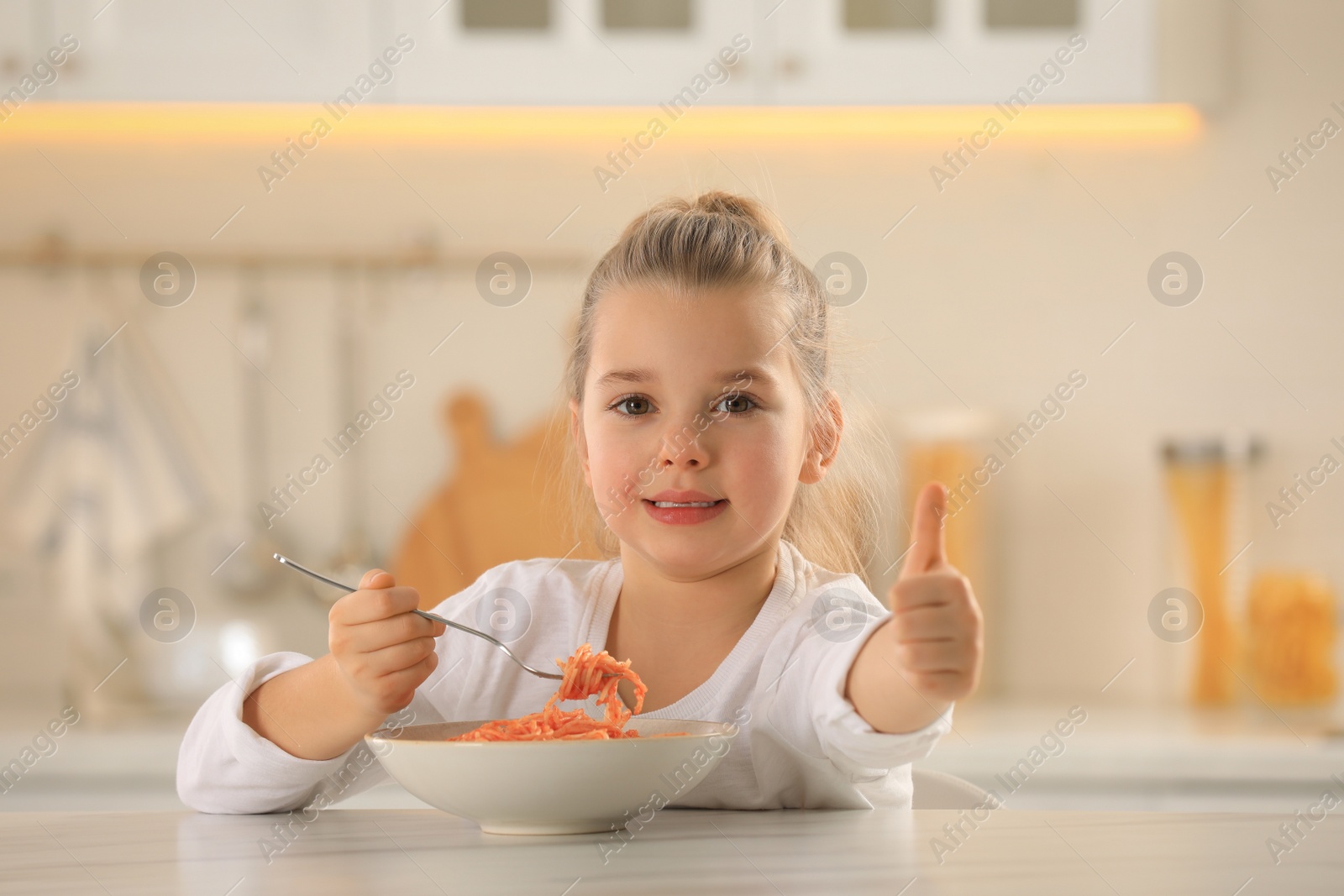 Photo of Cute little girl eating tasty pasta at table in kitchen