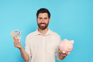 Happy man with money and ceramic piggy bank on light blue background