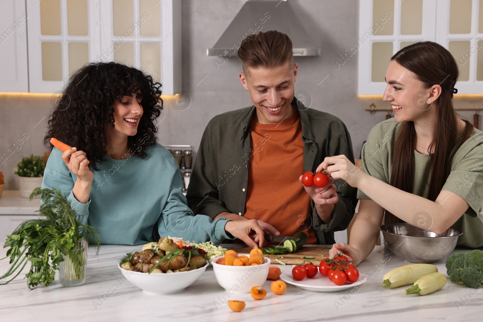 Photo of Friends cooking healthy vegetarian meal at white marble table in kitchen