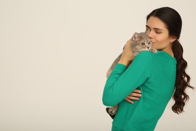 Young woman kissing her adorable cat on light background, space for text