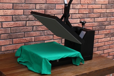 Photo of Heat press machine with t-shirt on wooden table near brick wall