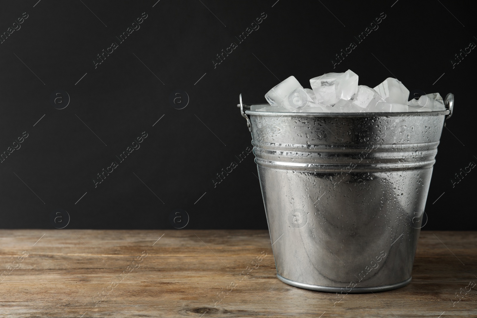 Photo of Metal bucket with ice cubes on wooden table. Space for text